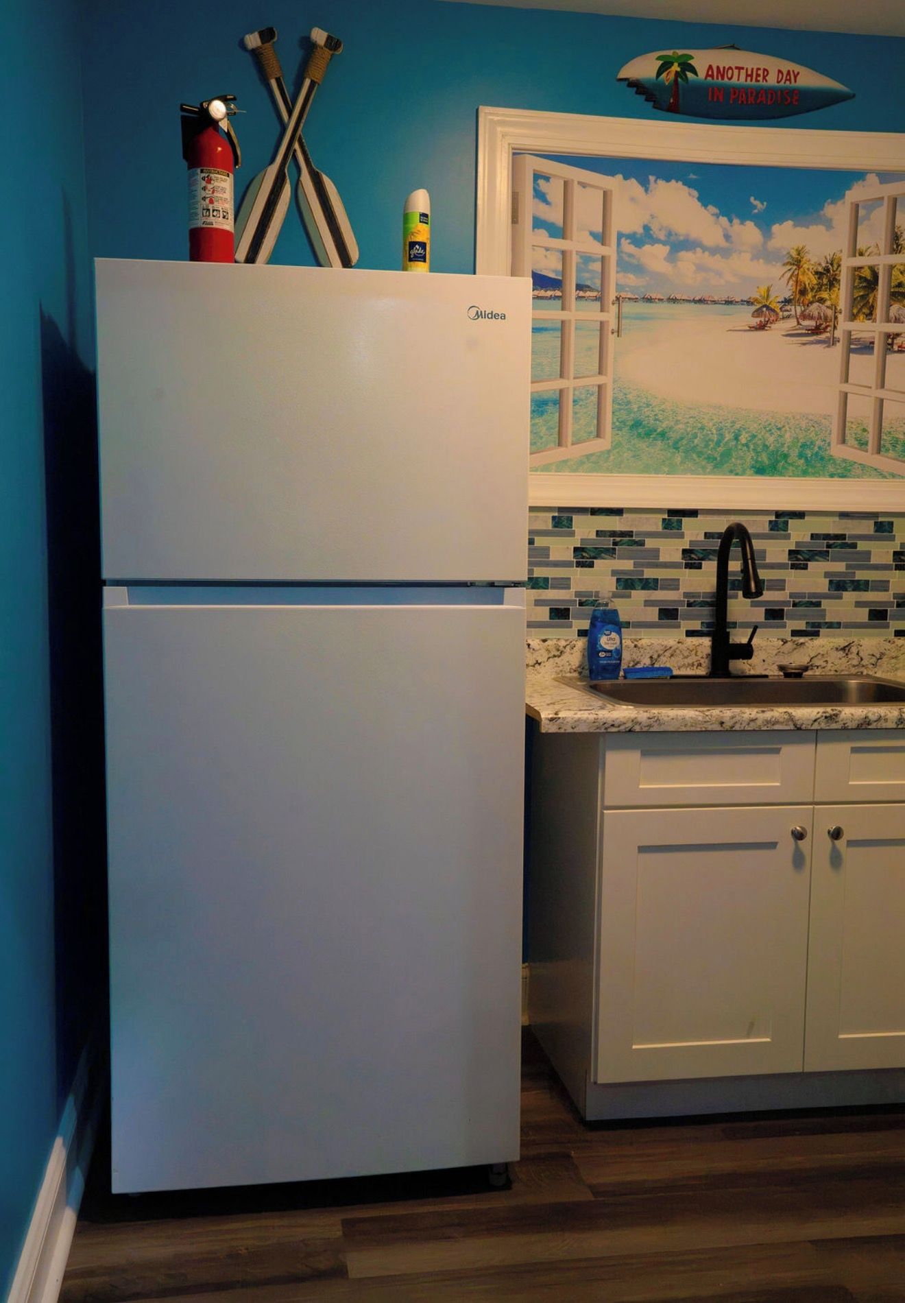 A kitchen with a white refrigerator, a tiled backsplash, a sink with a black faucet, and a window with a view of a beach.