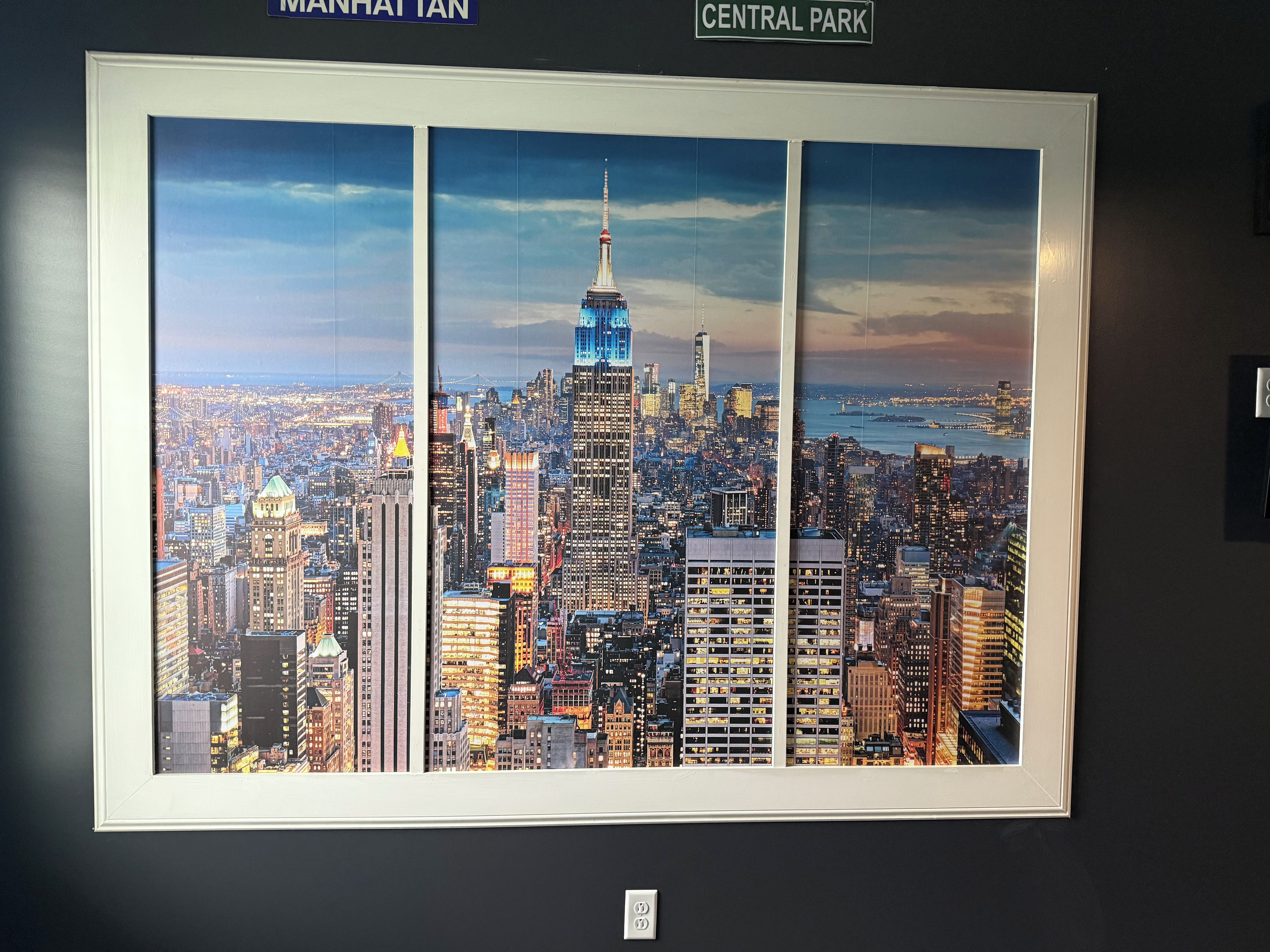 A framed cityscape artwork with a view of Manhattan's skyline, including the Empire State Building, mounted on a wall.