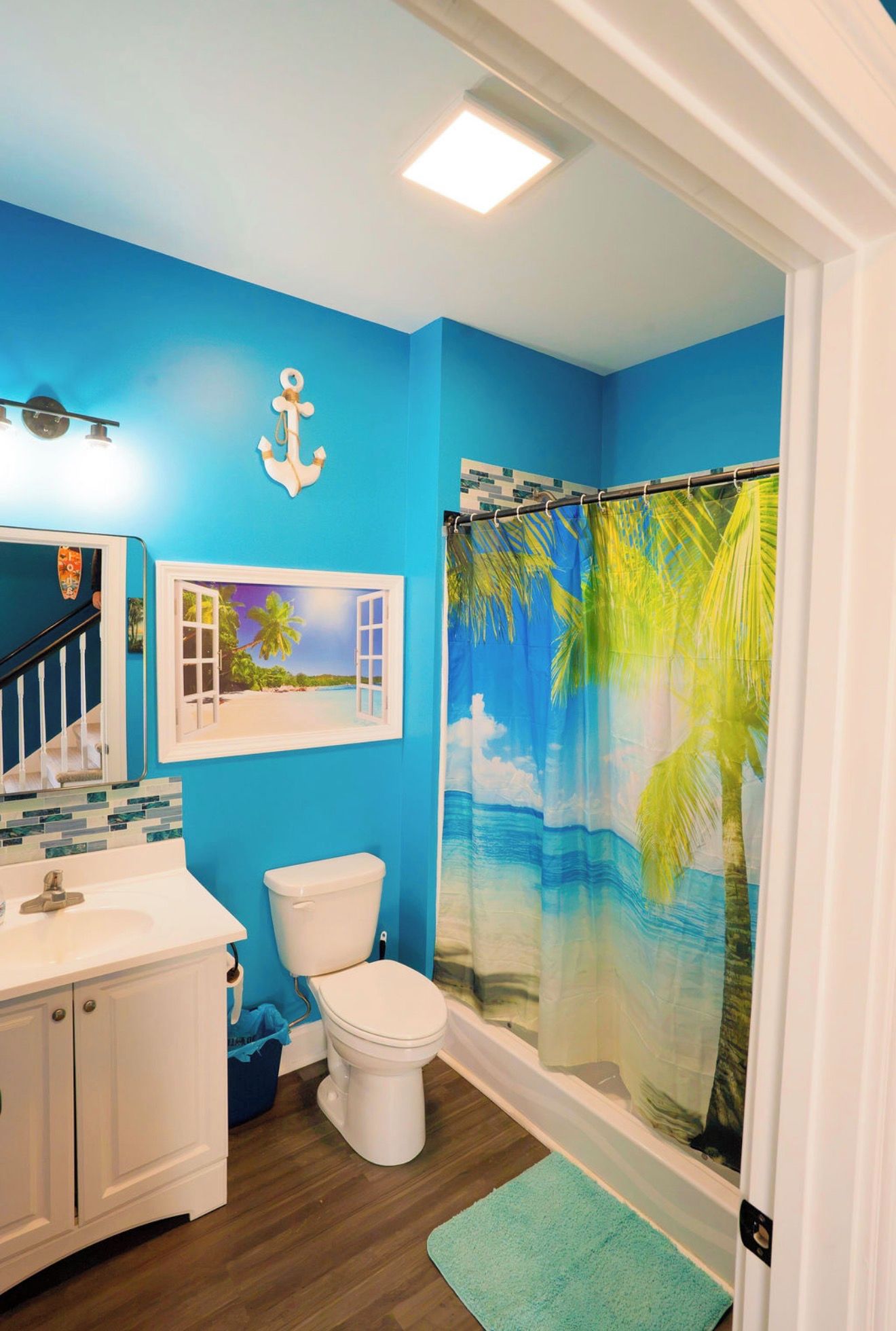 A bathroom with a blue wall, a white sink, a toilet, a shower curtain with a tropical design, and a small window with a beach scene.