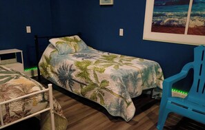 Un lit simple avec une couvre-lit à motifs tropicaux, une chaise bleue et une petite table blanche dans une pièce aux murs bleu foncé.