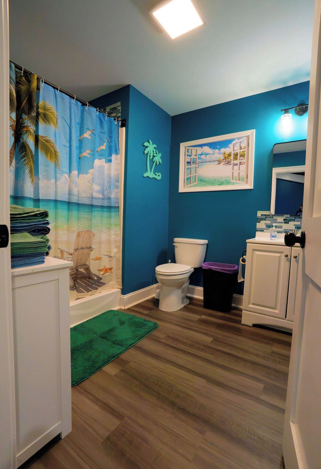 Ein Badezimmer mit blauer Wand, Toilette, einer Duschvorhang mit einer Strandansicht und einem grünen Teppich.