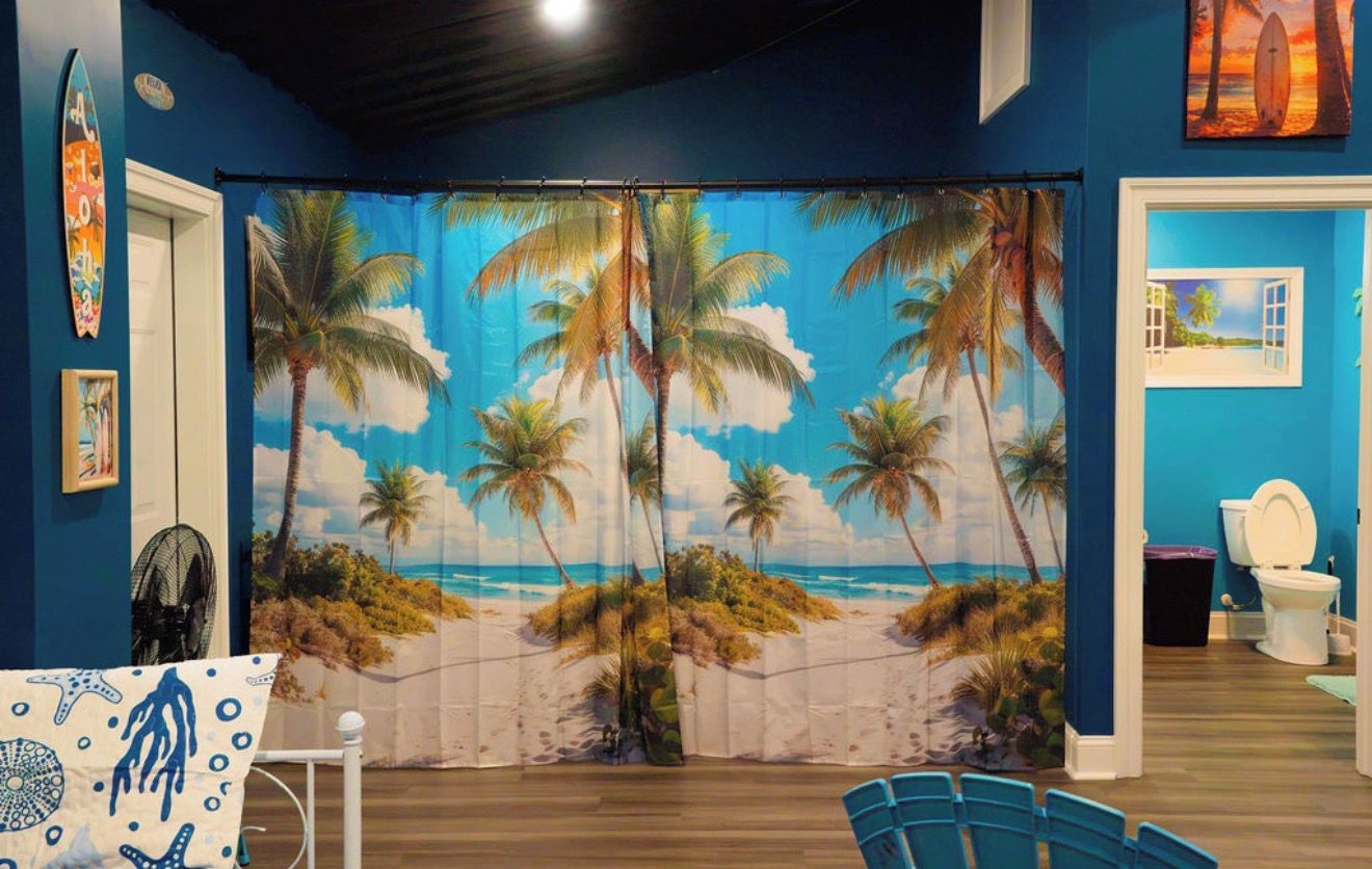 A room with a beach-themed curtain, a surfboard, and a window with a view of the beach.