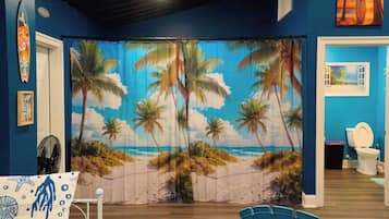 A room with a beach-themed curtain, a surfboard, and a window with a view of the beach.