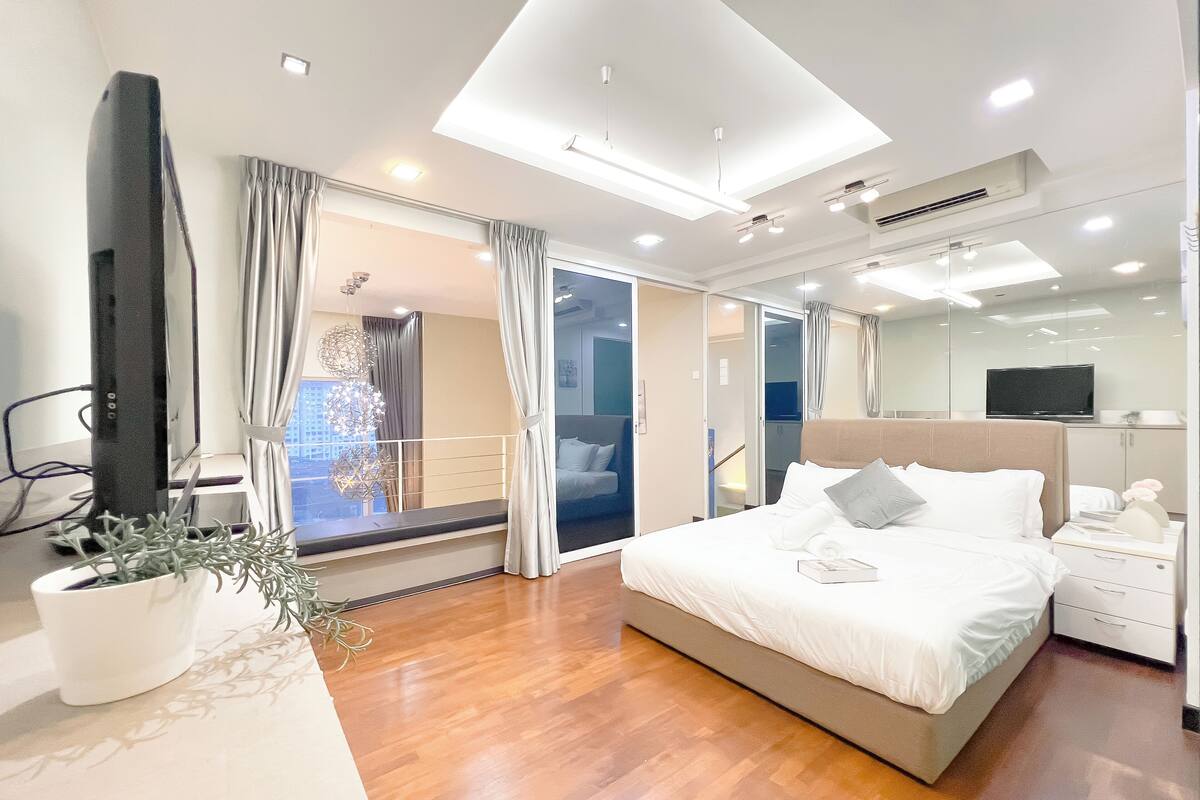 Una habitación de hotel moderna con una cama grande, un televisor, una planta en maceta y vistas de la ciudad por la ventana.