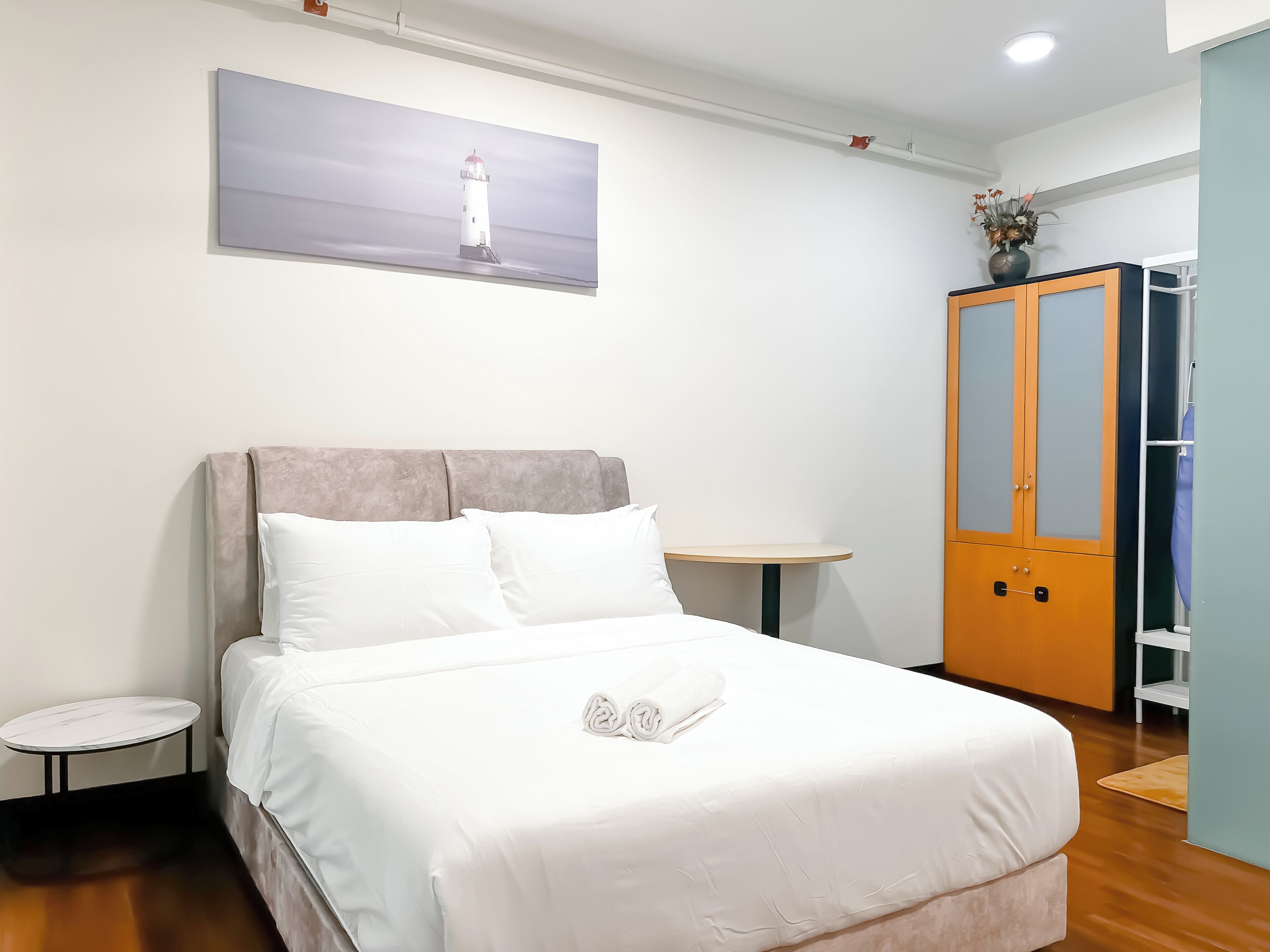 A neatly arranged hotel room with a bed, a small round table, a wardrobe, and a painting of a lighthouse on the wall.