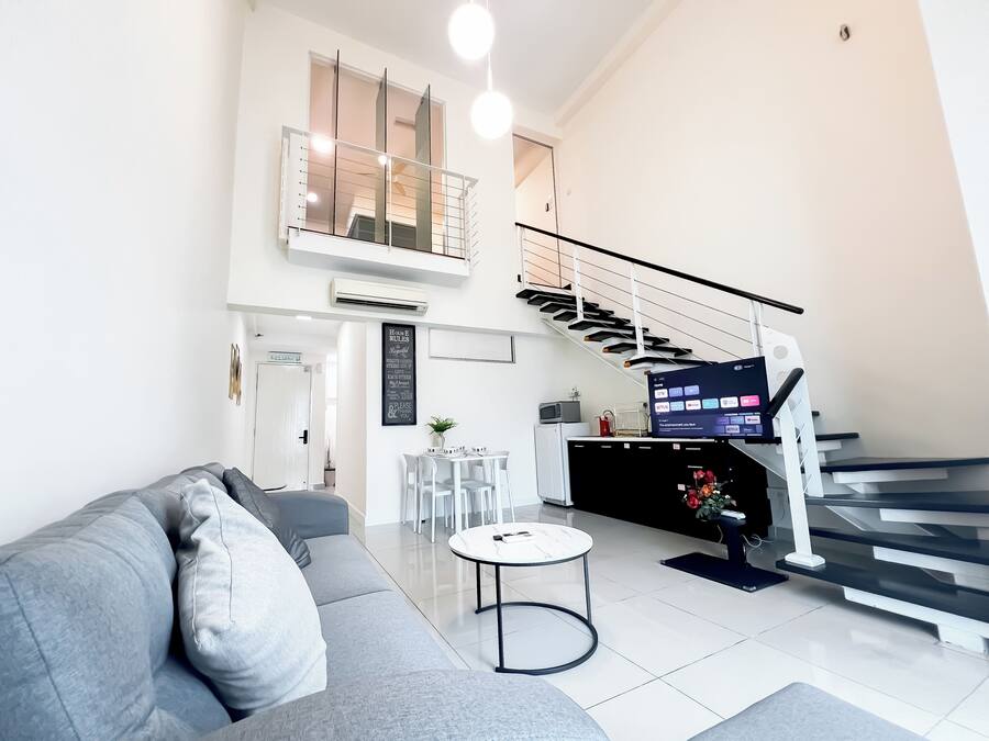 Una sala de estar moderna con un sofá gris, una mesita redonda, una escalera y una zona de cocina con un televisor.