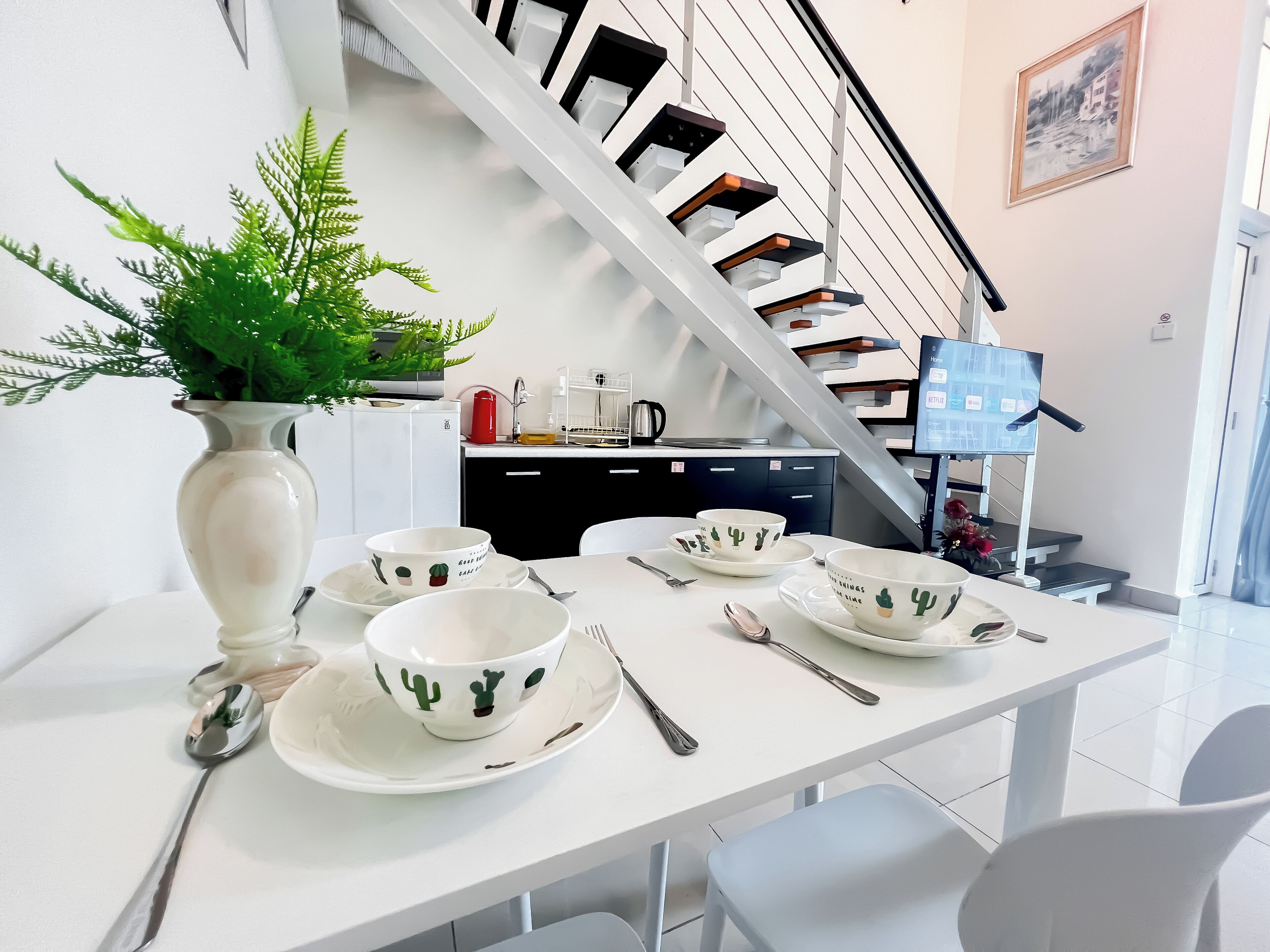 Un coin repas avec une table blanche dressée pour quatre personnes, une plante en pot et une cuisine en arrière-plan.