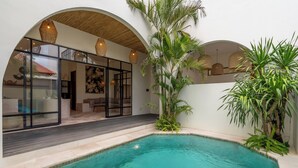 A swimming pool with a tiled deck, surrounded by indoor seating areas and plants. - Sofia Villa by Betterplace (Canggu)