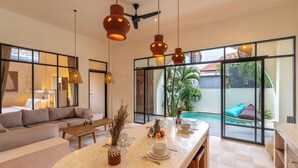 A modern hotel room with a large window, a sofa, a dining area with a table set for two, and a ceiling fan. - Sofia Villa by Betterplace (Canggu)