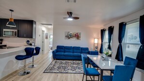 A modern living room with a blue sofa, a dining table with chairs, a kitchen area with dark cabinets, and a ceiling fan.