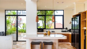 A modern kitchen with a large island, wooden stools, a black refrigerator, and a view of a garden through large windows. - Leo Villa by Betterplace (Canggu)