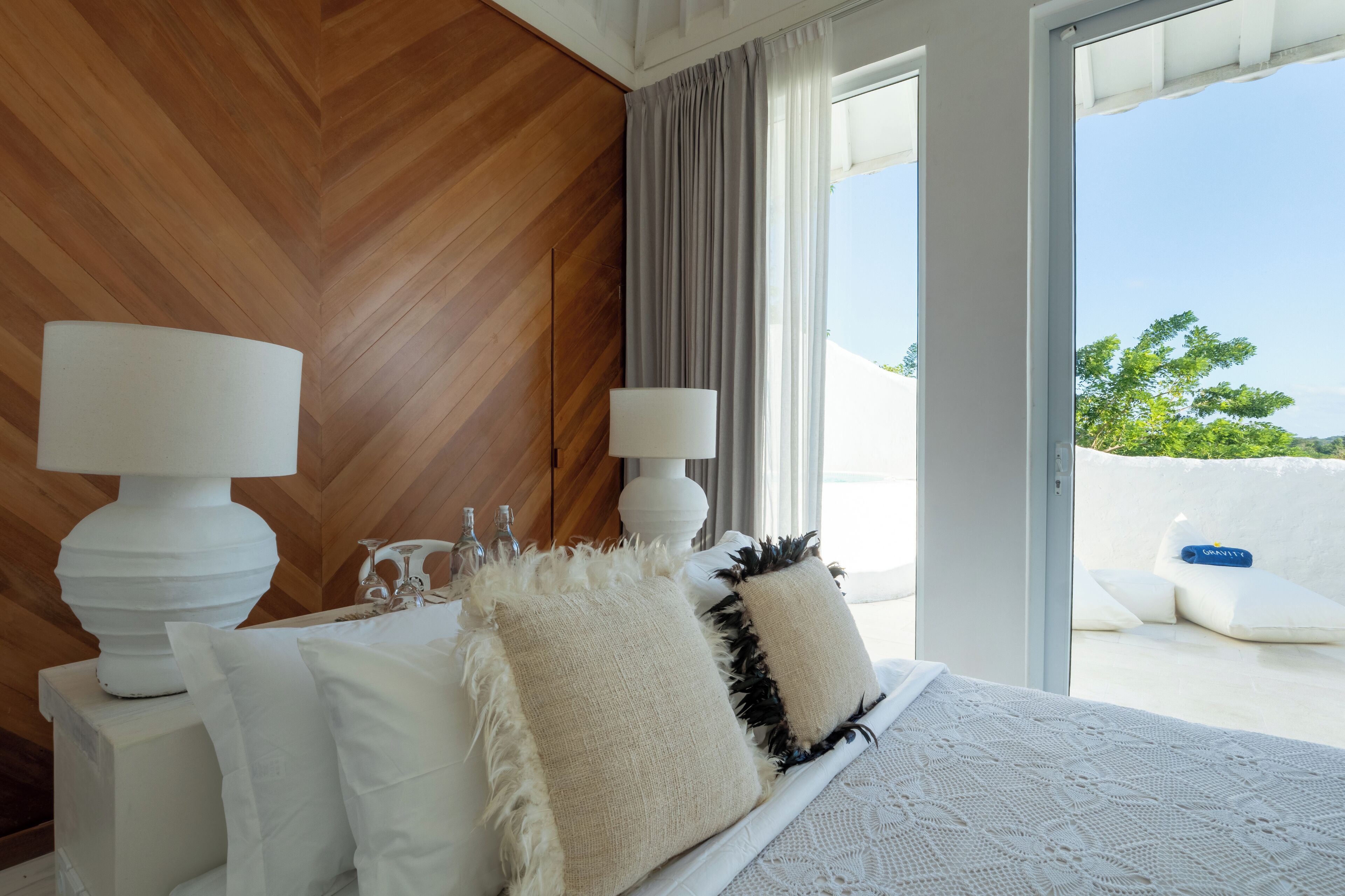 A bedroom with a wooden headboard, two bedside lamps, a large bed with white bedding, and a view of a balcony with outdoor furniture.