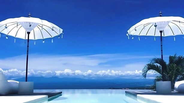 Ein Poolbereich mit zwei weißen Sonnenschirmen, blauem Himmel und Blick aufs Meer.
