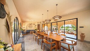 A spacious dining area with a long wooden table and chairs, large windows, and a view of the outdoors. - Cabana Kedungu by Ini Vie Hospitality  (Tabanan)