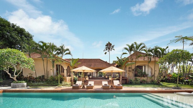 A swimming pool with lounge chairs and umbrellas, surrounded by palm trees and a building with a tiled roof. - Cabana Kedungu by Ini Vie Hospitality  (Tabanan)
