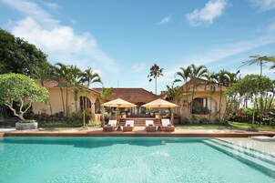 A swimming pool with lounge chairs and umbrellas, surrounded by palm trees and a building with a tiled roof.