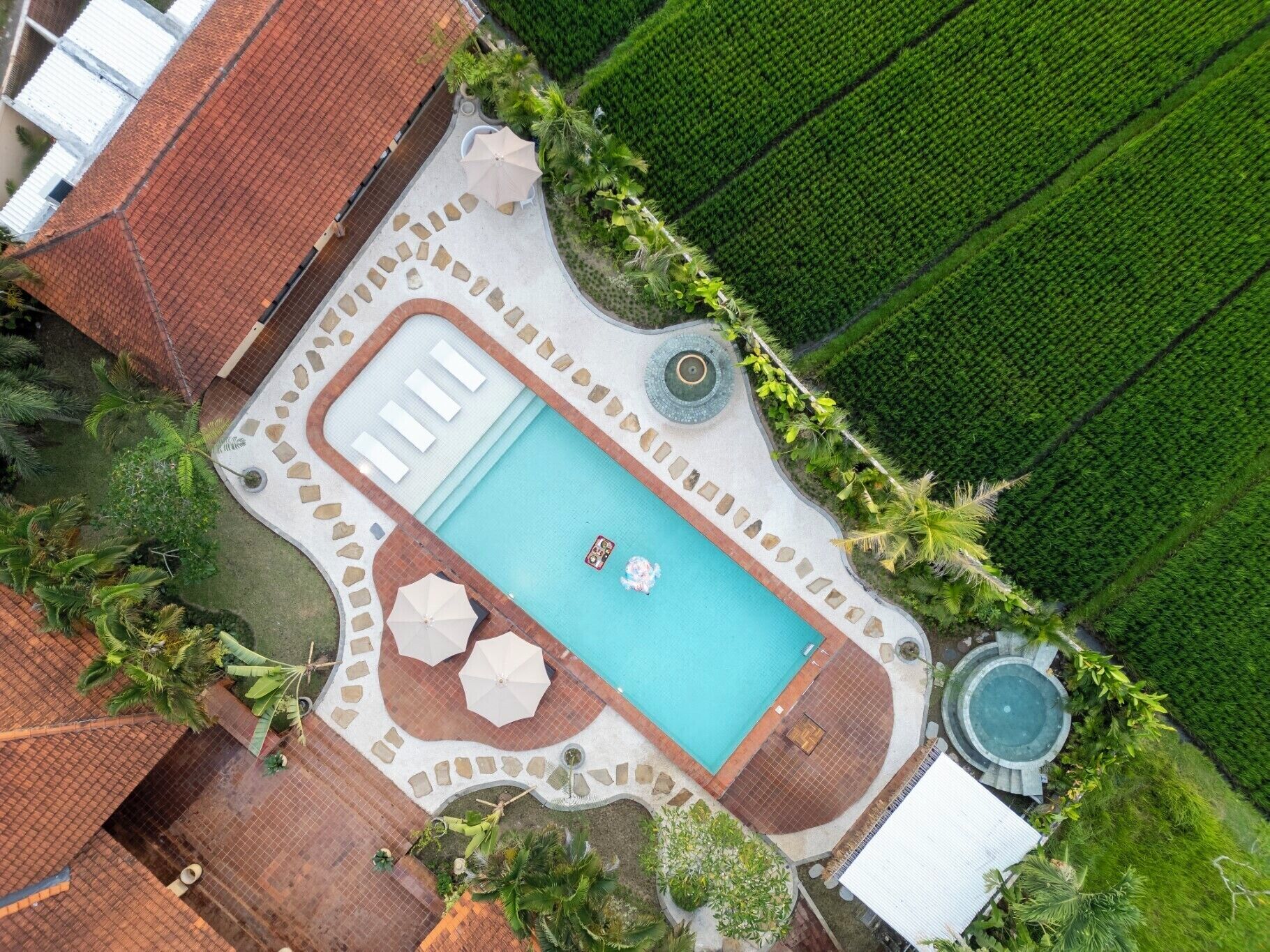 An aerial view of a swimming pool area with a rectangular pool, surrounded by a stone deck, and adjacent to a lush green field.