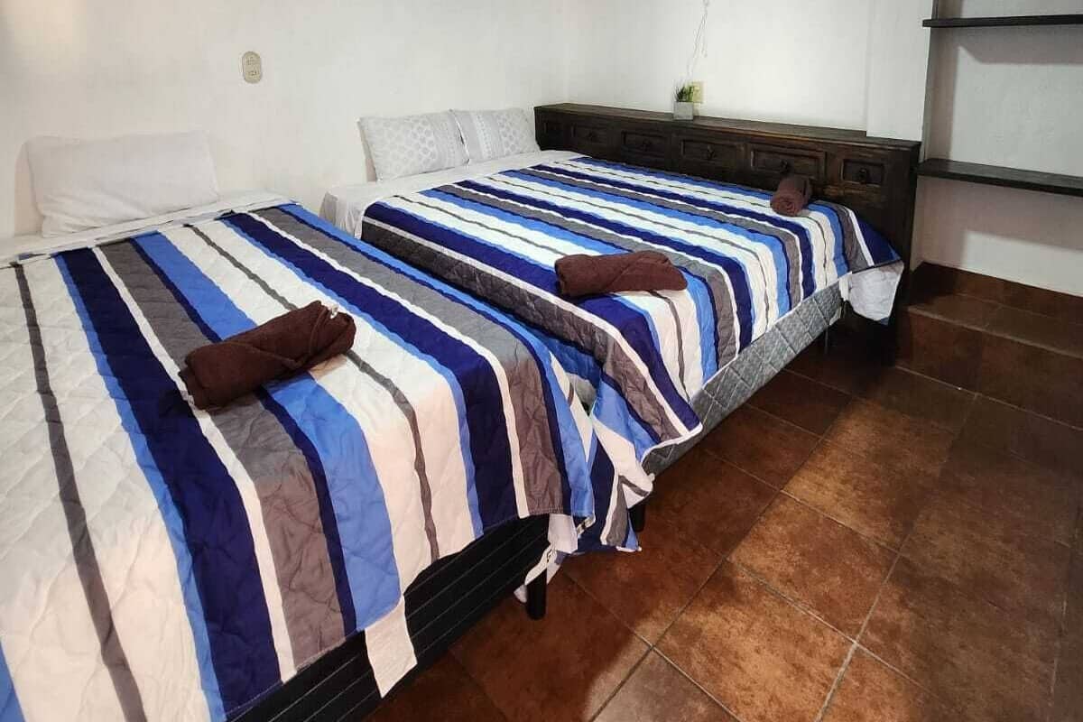 A room with two beds, a brick-patterned ceiling, a wall fan, and a small kitchenette.