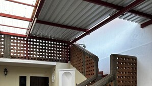 Un espacio al aire libre techado con una escalera que conduce a un nivel superior, suelo de piedra y estructura de techo metálico.