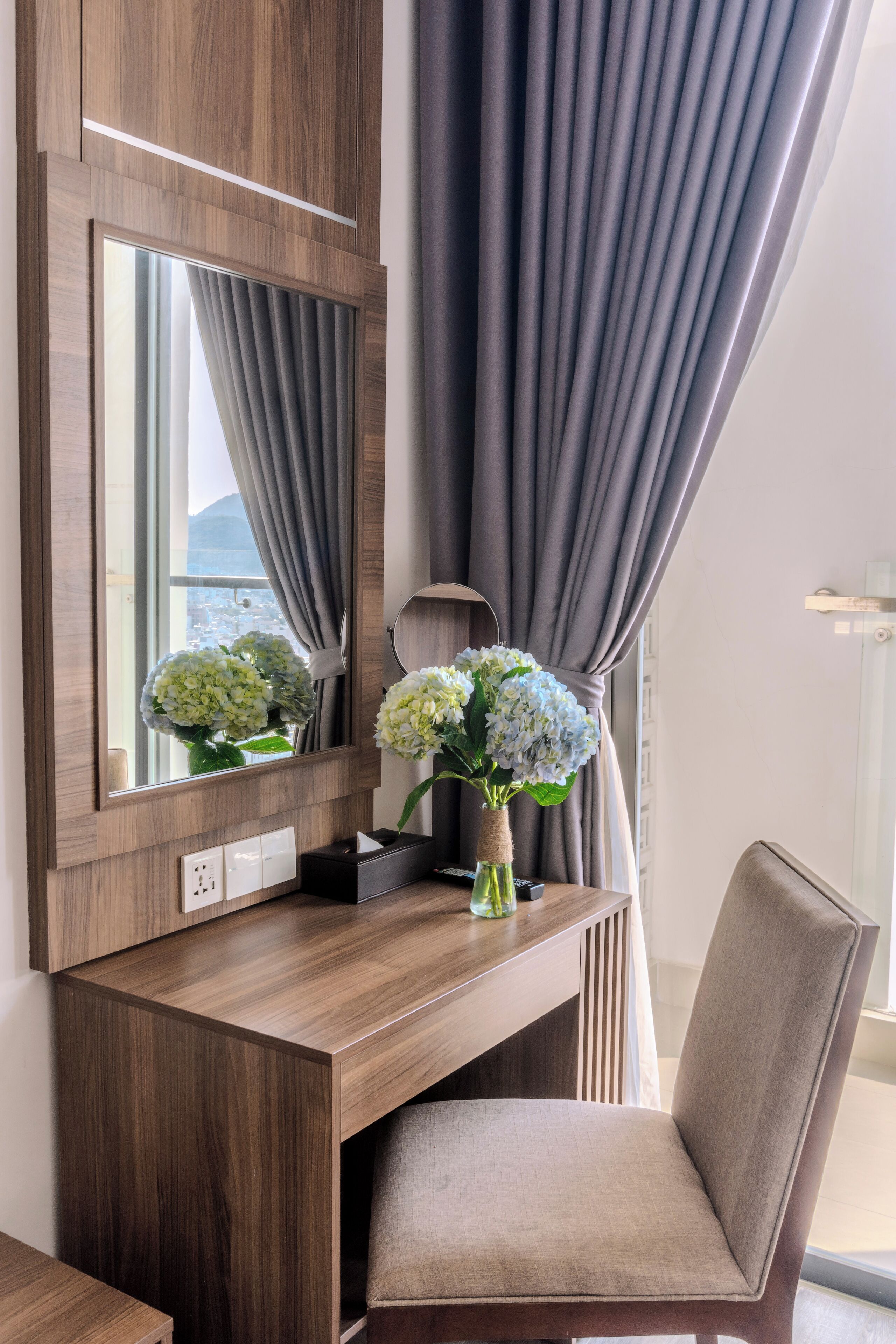 A wooden desk with a mirror, a vase with flowers, and a chair.