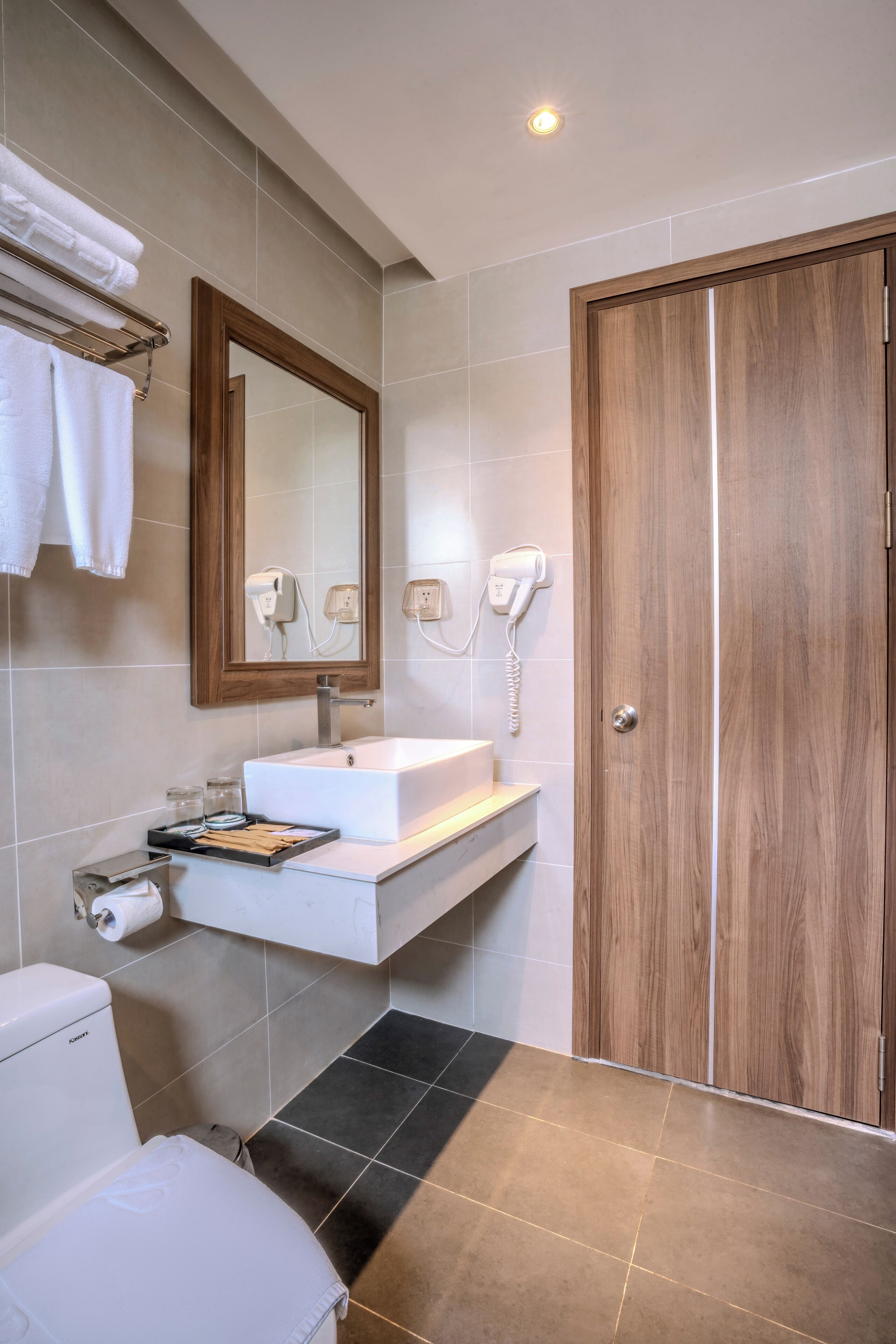 A modern bathroom with a white sink, a wooden door, and a mirror with a light switch.