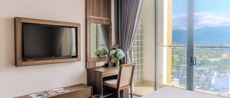 A hotel room with a large TV, a desk, a chair, a bed, and a view of a cityscape through a large window.