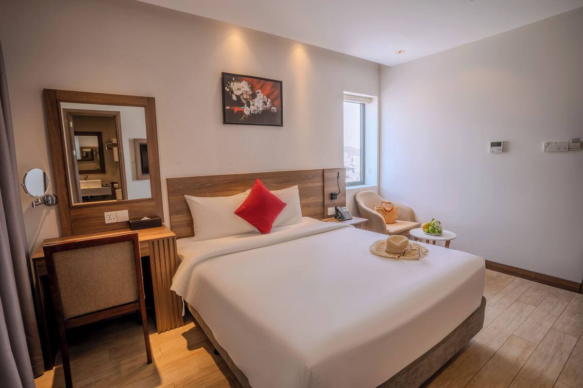 Ein Hotelzimmer mit einem großen Bett, einem Schreibtisch mit Stuhl, einem kleinen Tisch mit Korb, einem Spiegel und einem Fenster.