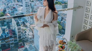 Eine Frau steht an einem Balkon mit Blick auf die Stadt, eine Tasse haltend. Vor ihr steht ein kleiner Tisch mit einer Speiseplatte und einem Blumentopf.