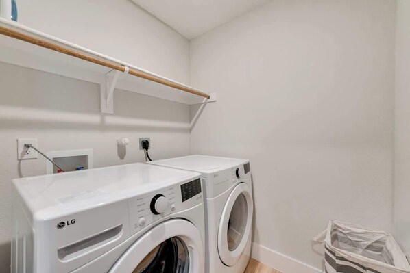 Una lavanderia con due lavatrici LG, uno scaffale in legno, un muro bianco e un cestino.