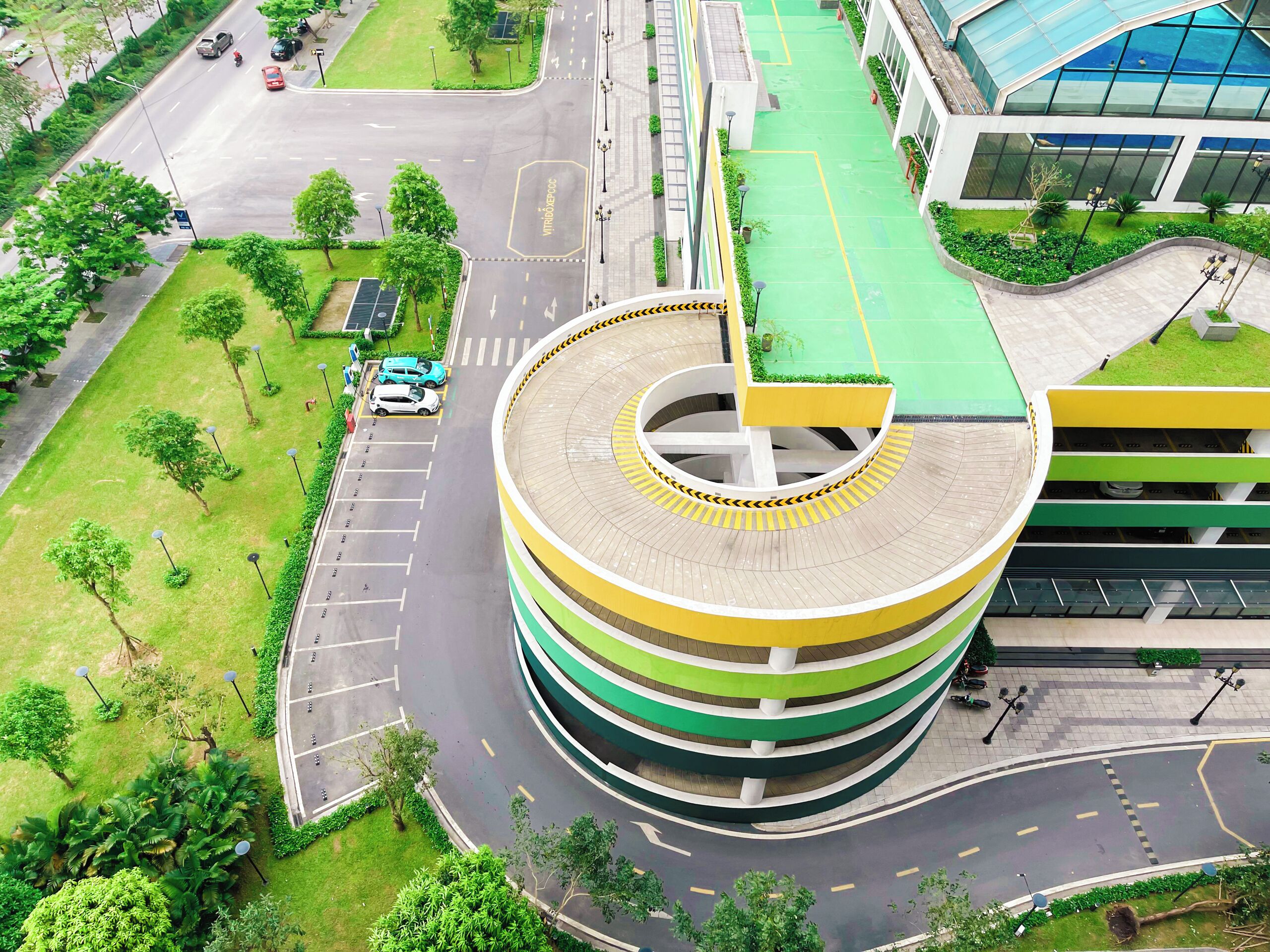 Una estructura de estacionamiento de varios niveles con diseño circular, rodeada de áreas verdes y una carretera con vehículos.