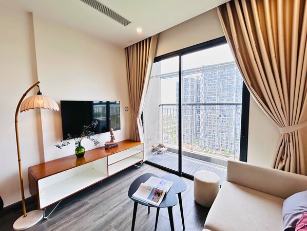 A modern hotel room with a flat-screen TV, a wooden console table, a sofa, a small round table with a magazine, and a large window with a view of high-rise buildings.