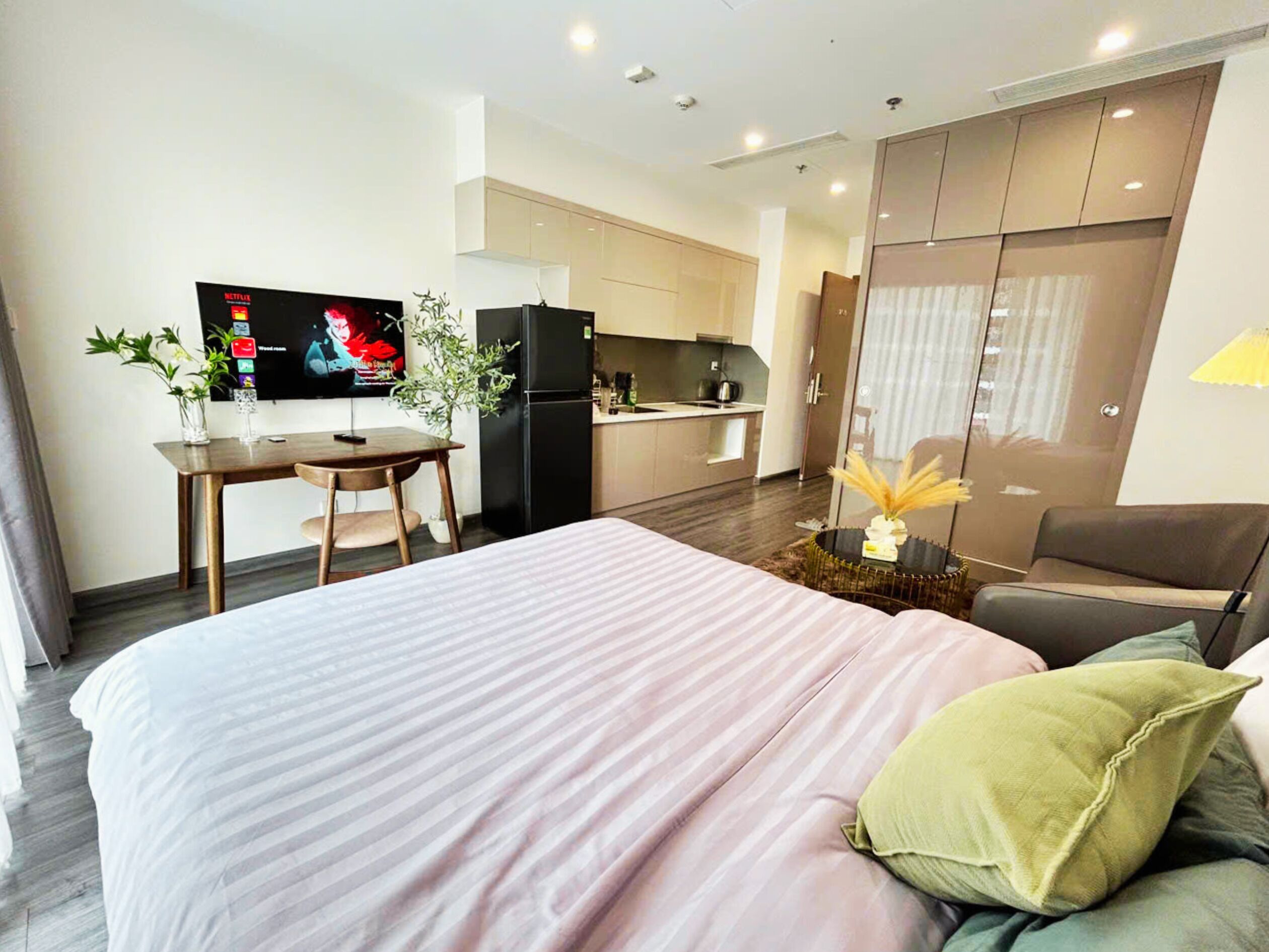 A modern hotel room with a large bed, a desk with a TV, a kitchenette, and a separate seating area.