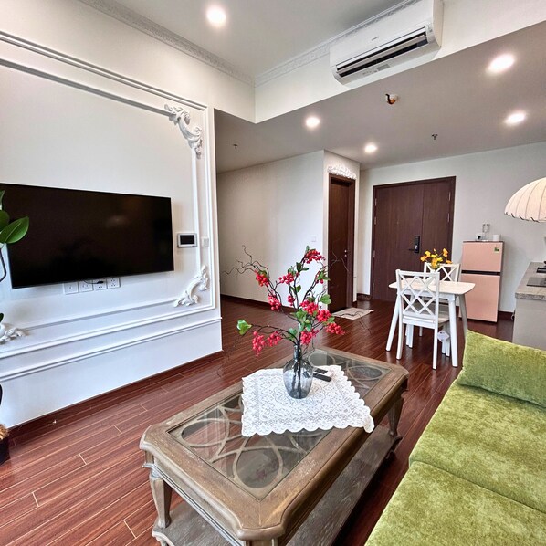 A living room with a flat-screen TV mounted on the wall, a glass coffee table with a lace doily, a vase with red flowers, a green sofa, and a dining area with white chairs and a table.