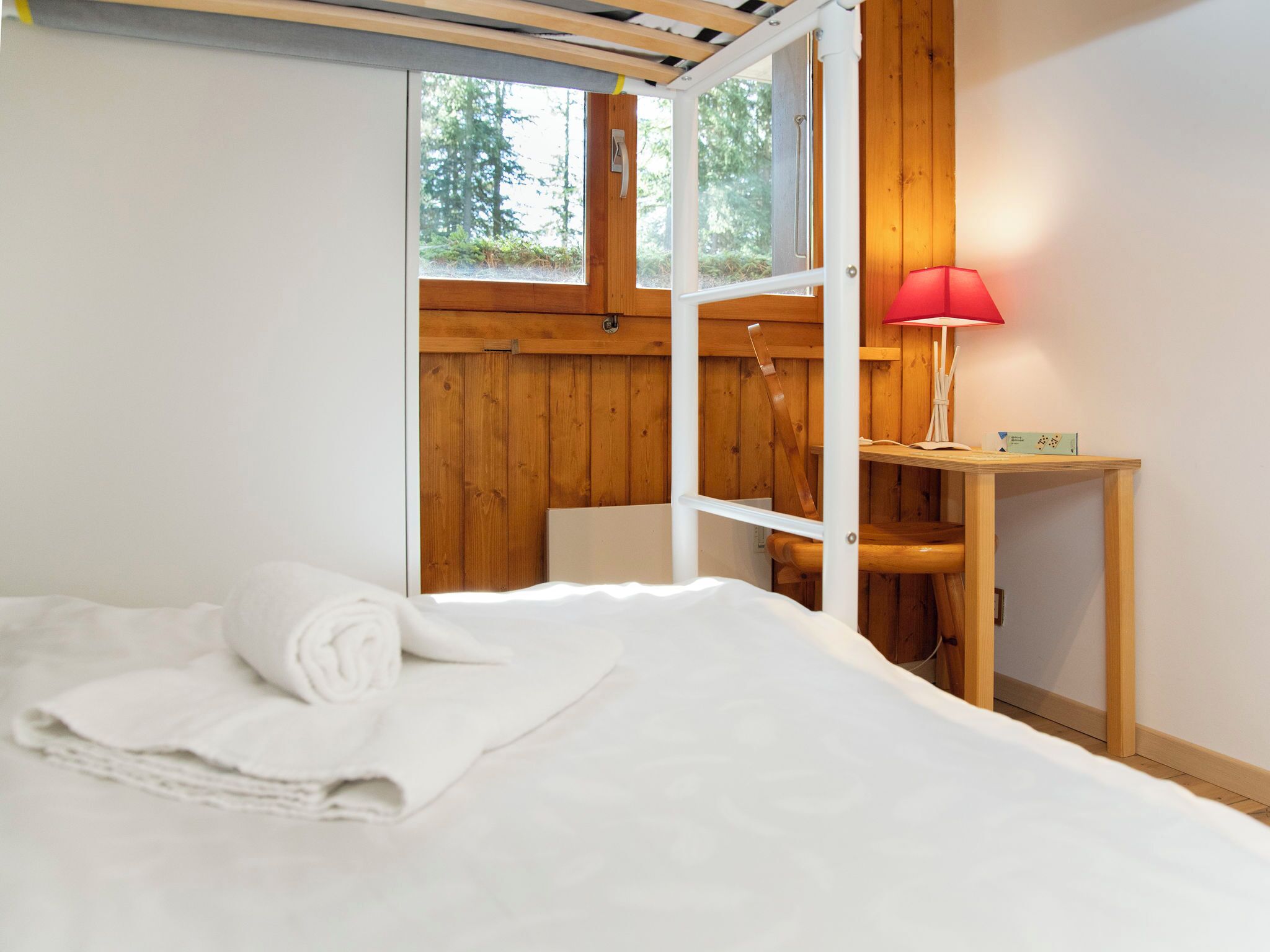 A bedroom with a bed, a wooden desk, a red lamp, and a window with a view of trees.