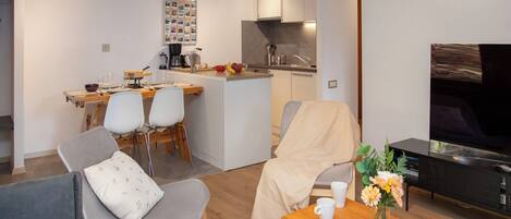 Una sala de estar moderna con un sofá gris, una mesa de centro de madera y un televisor. Cuenta con una zona de cocina con una mesa de comedor y sillas.