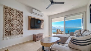 A modern living room with a large window, a ceiling fan, a flat-screen TV, and a cozy seating area with a view of the ocean.