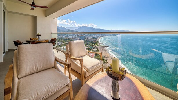 A balcony with a view of the ocean and cityscape, furnished with chairs and a table, and a ceiling fan.