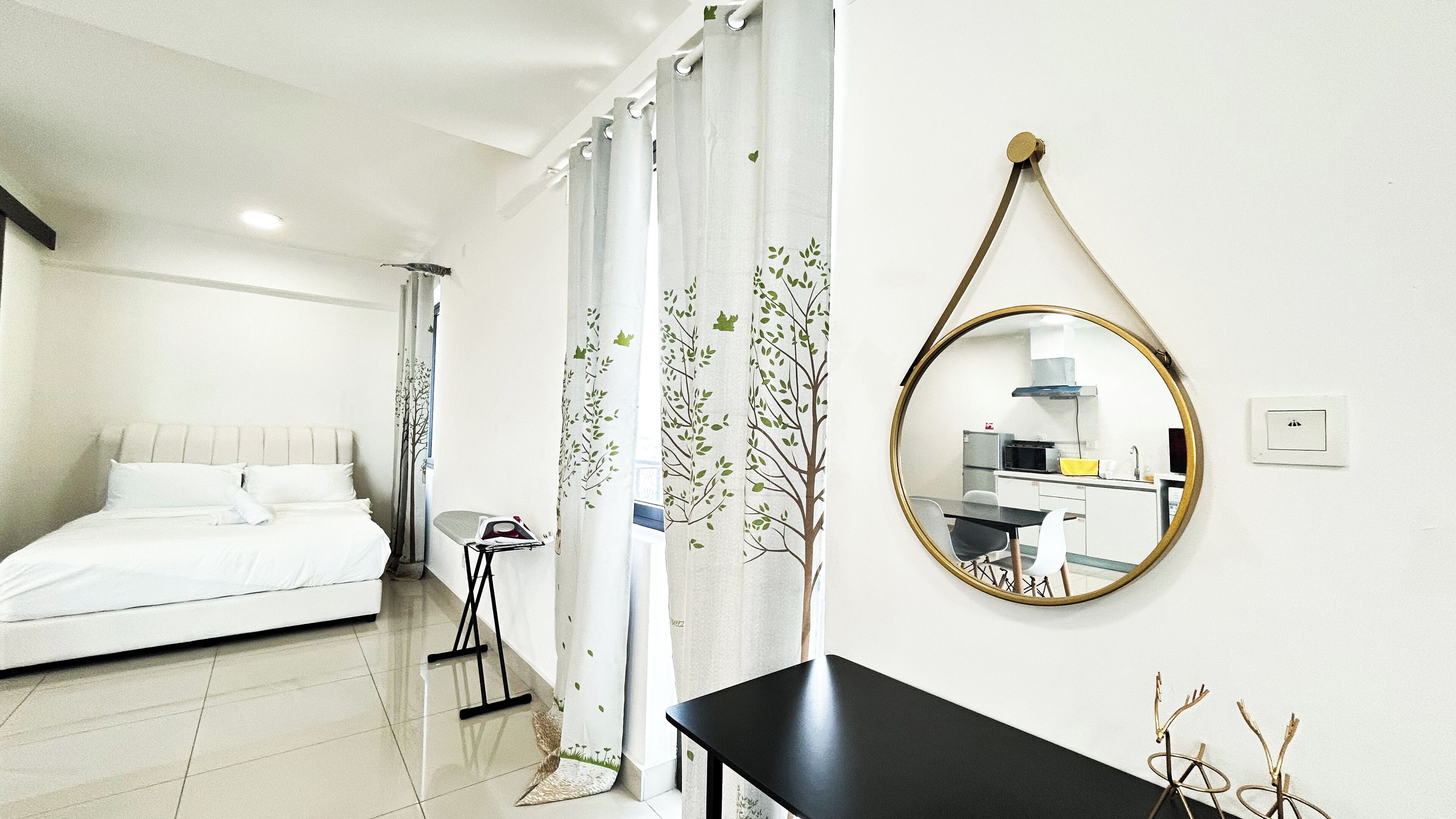 A bedroom with a bed, a small table, a mirror, and a view of a kitchen through a window.