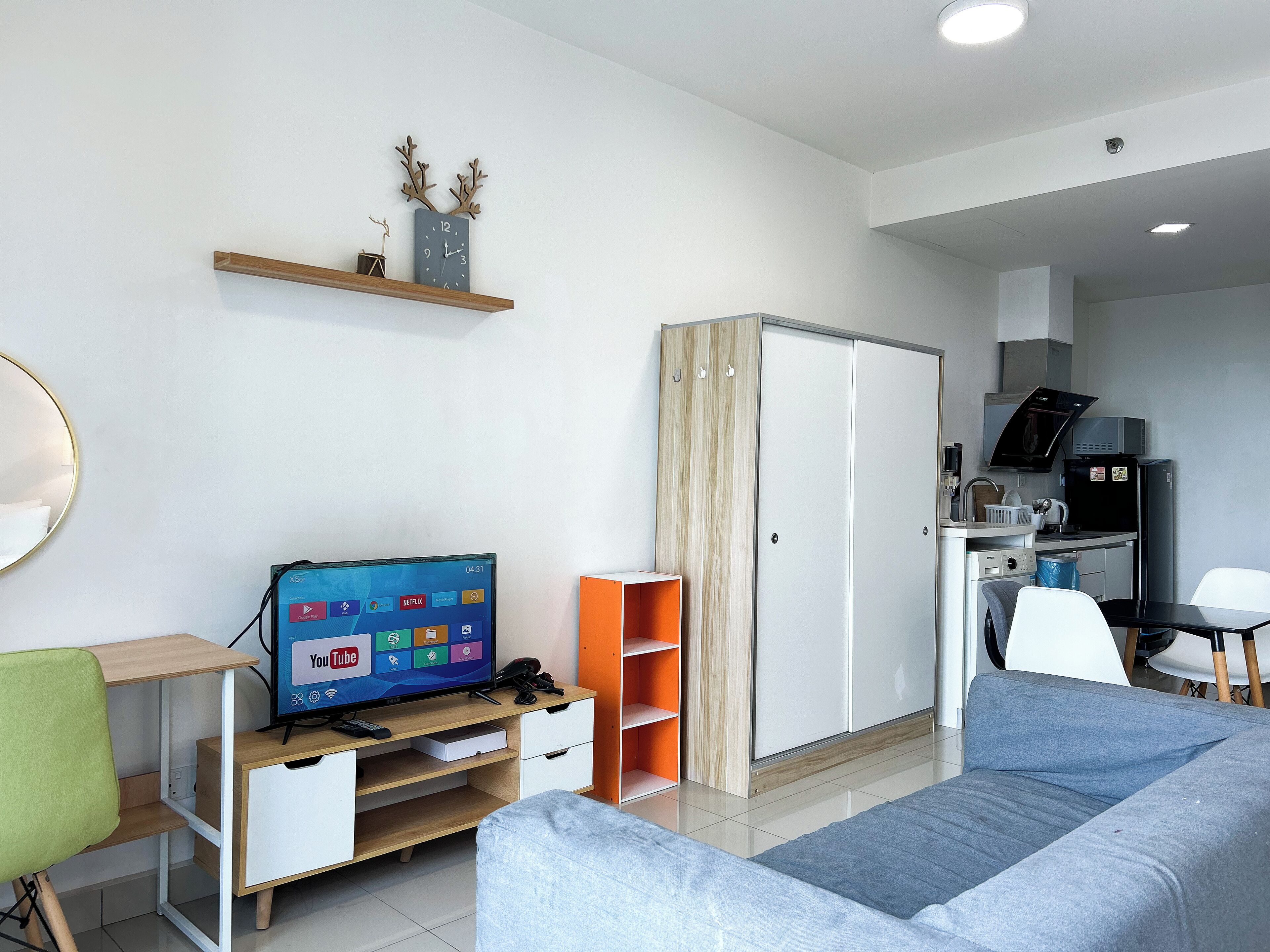 A modern, well-lit room with a flat-screen TV, a wooden desk, a sofa, a wardrobe, and a kitchenette.