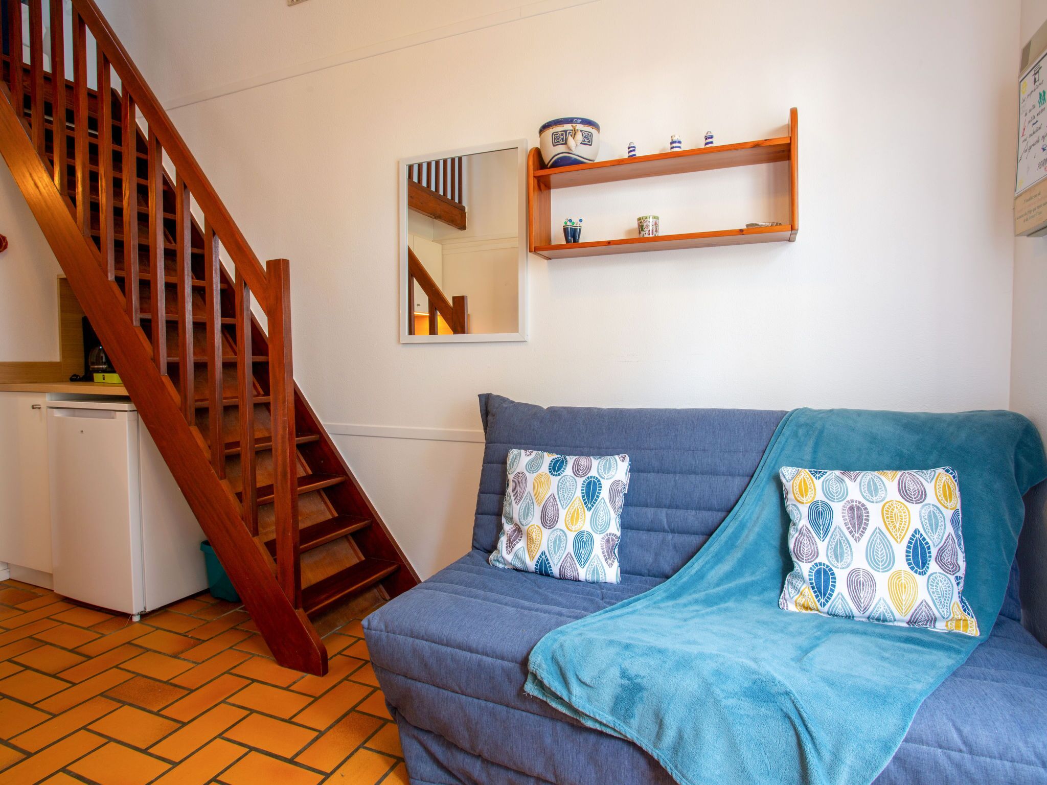 A room with a wooden staircase, a blue sofa with patterned cushions, a small white fridge, and a shelf with decorative items.