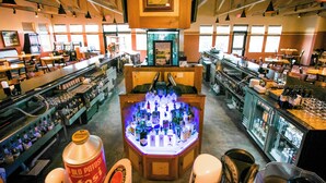 Zona de bar con un mostrador central lleno de botellas, rodeada de asientos y estanterías con más botellas.