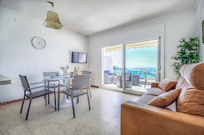 A modern living room with a balcony, a dining area, and a TV. - Beachfront Apartment in Platja Almadrava (Roses)