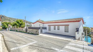 A white building with a red-tiled roof, a stone wall, and a paved road with a pedestrian crossing. - Beachfront Apartment in Platja Almadrava (Roses)
