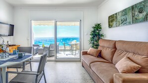 A modern living room with a sofa, dining table, and chairs, a TV, and a view of the ocean through large sliding doors. - Beachfront Apartment in Platja Almadrava (Roses)