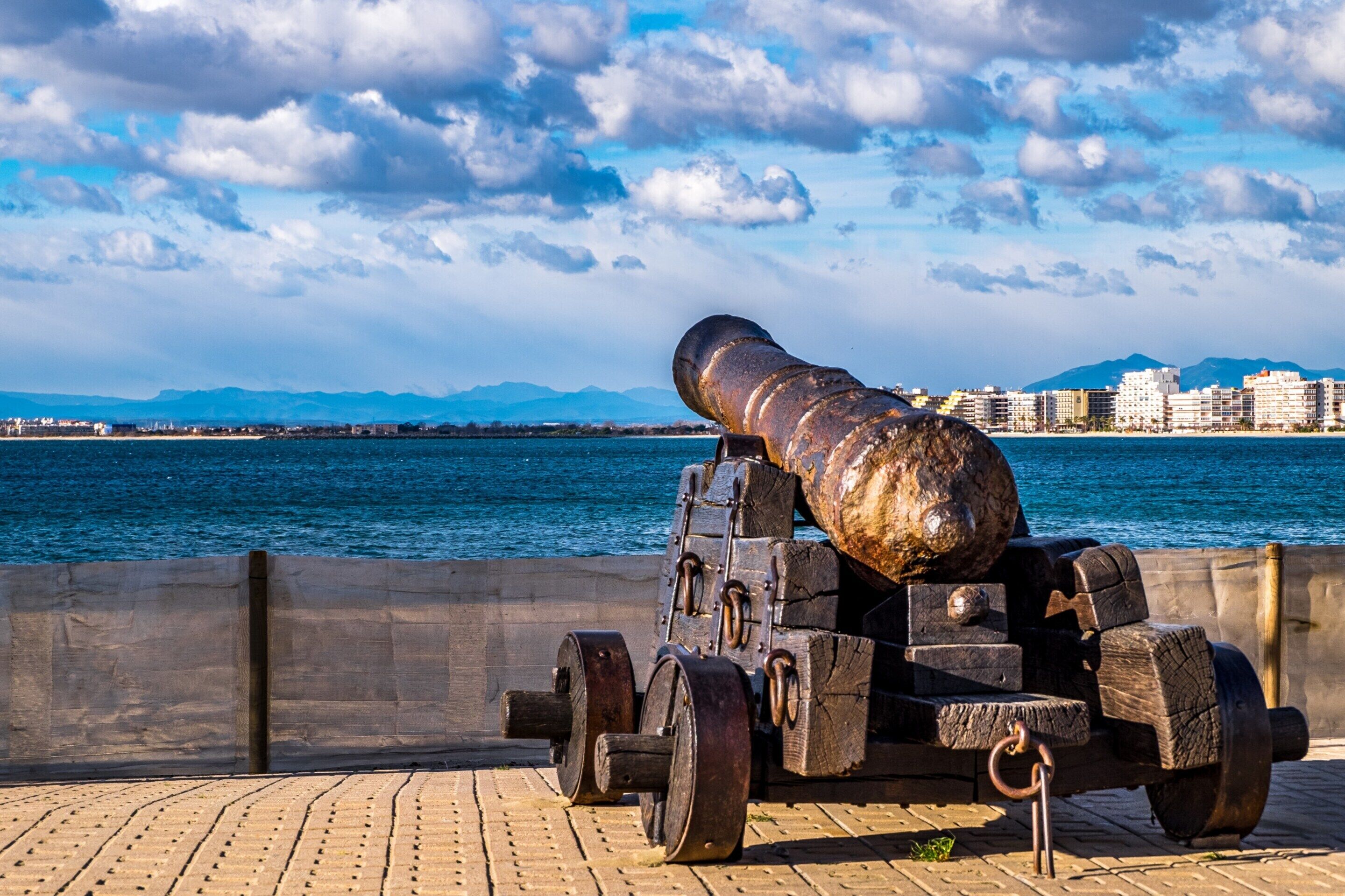A cannon on a wheeled platform by a waterfront with buildings and mountains in the background.