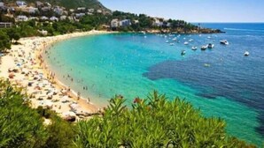 A coastal beach with clear turquoise waters, a sandy shore, and a backdrop of buildings and greenery. - Beachfront Apartment in Platja Almadrava (Roses)