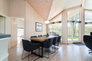 A modern interior with a wooden ceiling, a dining table with chairs, and large windows offering a view of the outdoors.