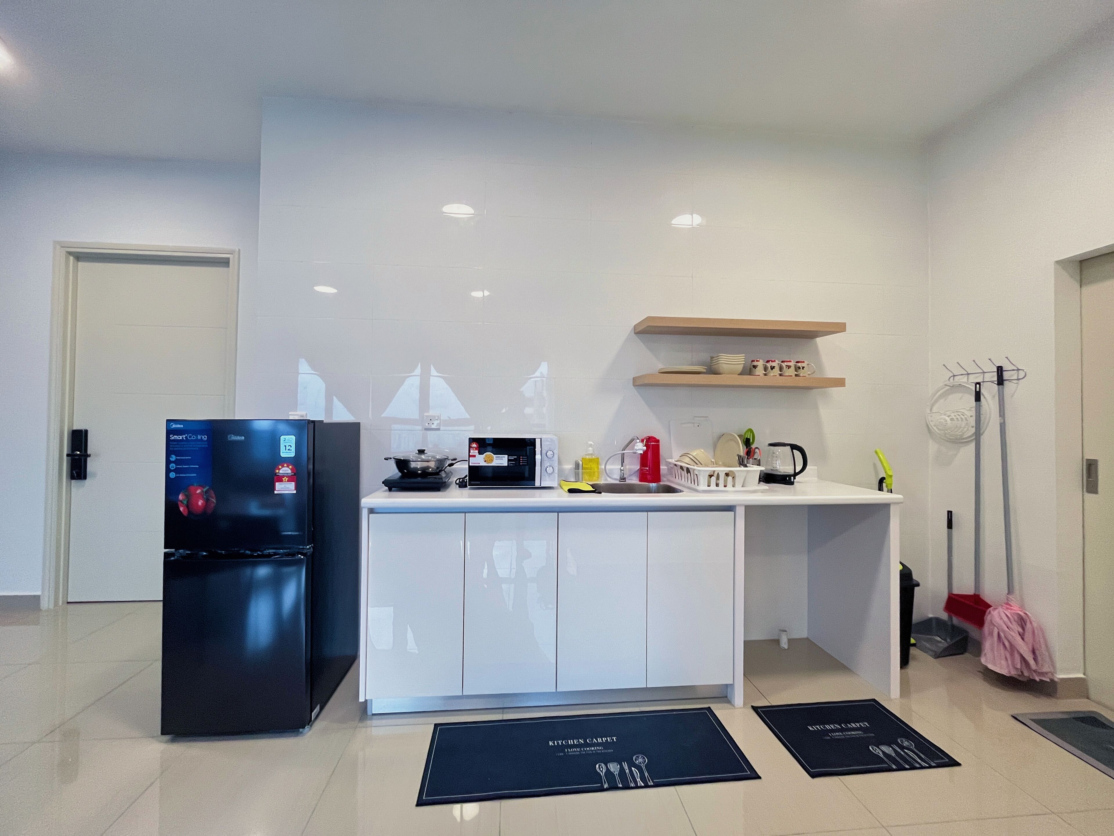 Een moderne keuken met een zwarte koelkast, witte kastjes, een magnetron en een spoelbak. Op de vloer liggen twee zwarte matten, waarvan één met de tekst 'Keuken Tapijt' en de andere 'Air Conditioner'.