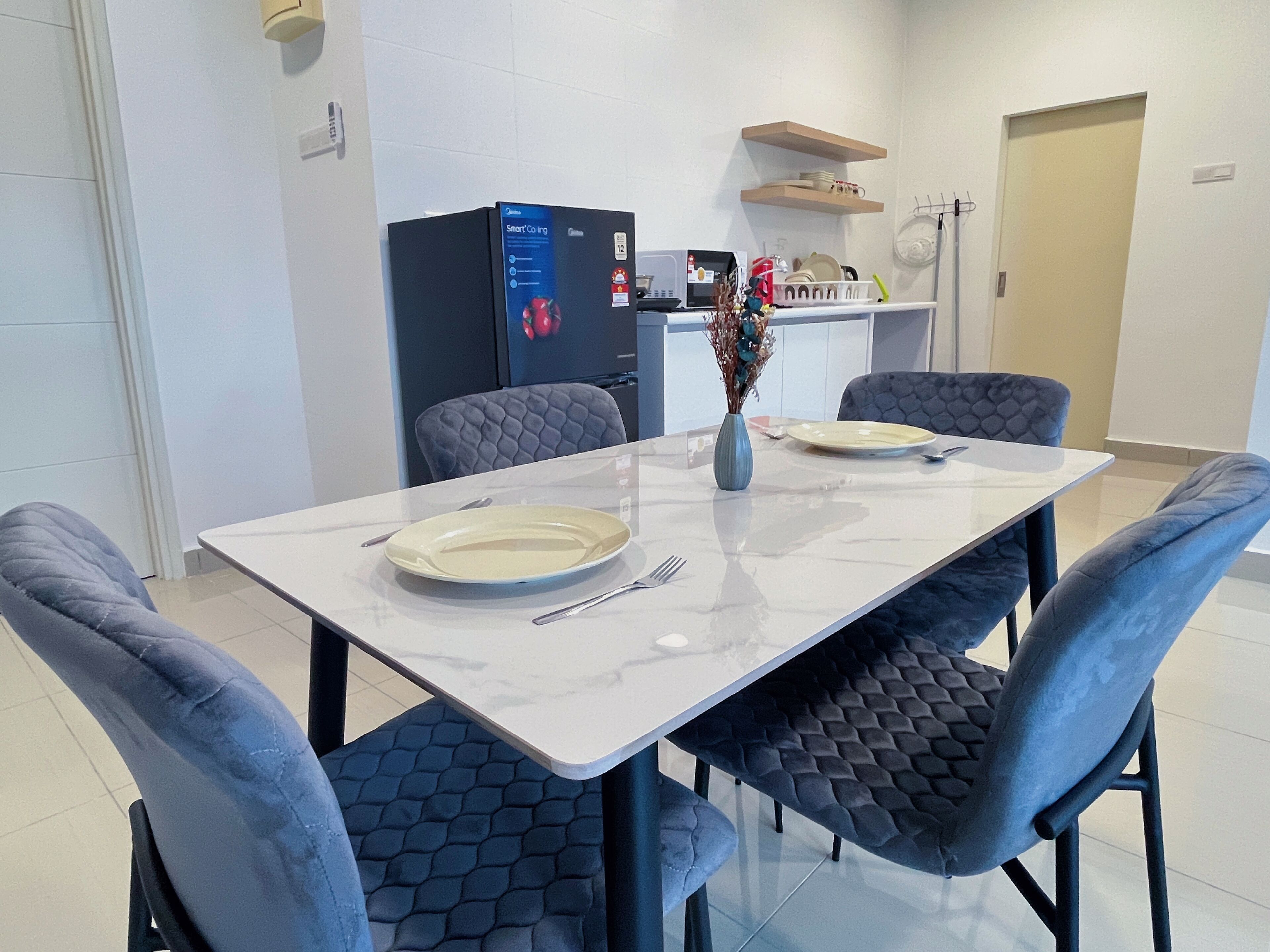 A modern dining area with a white table, two grey upholstered chairs, a vase with dried flowers, and a microwave in the background.