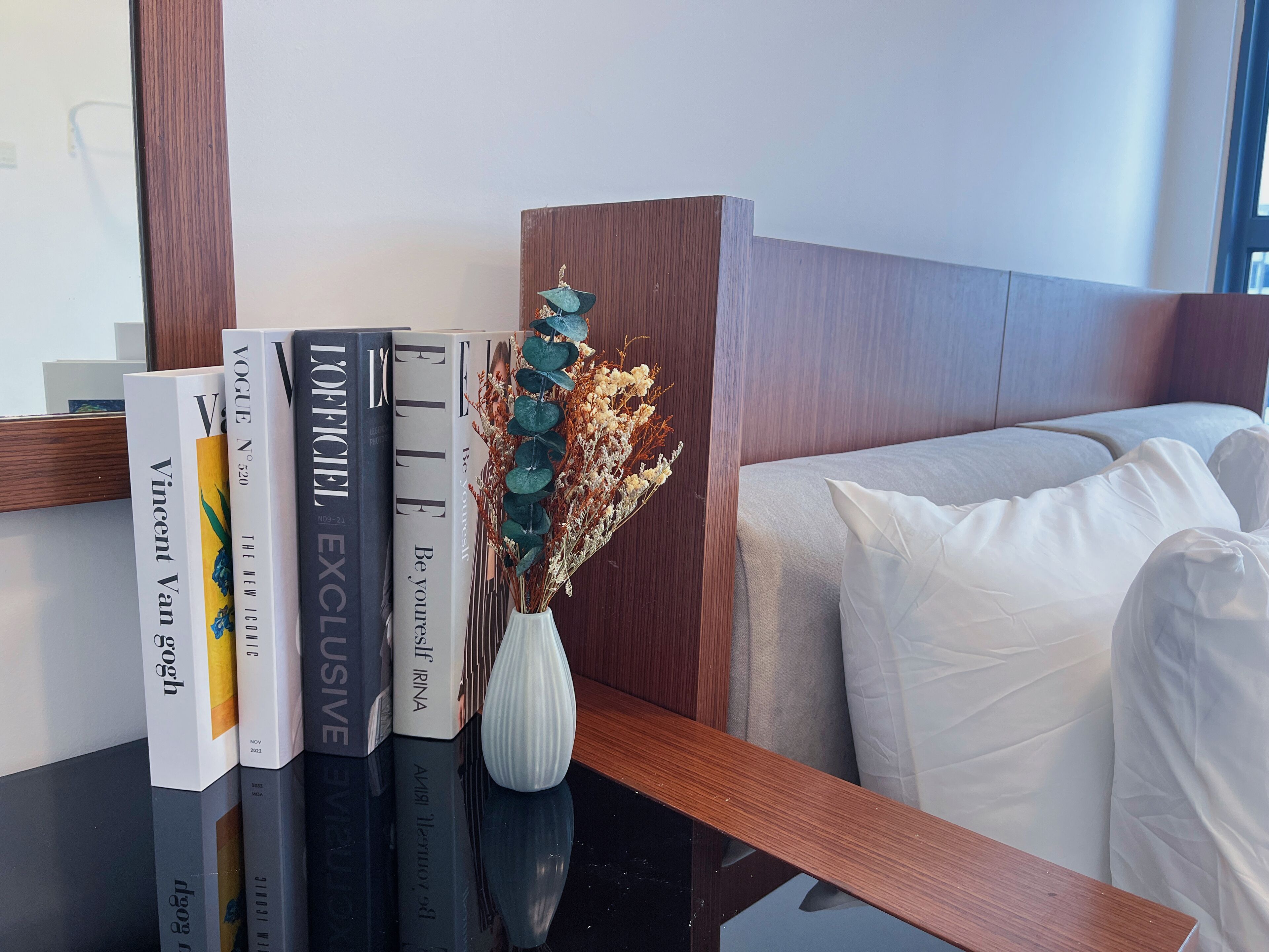 Una estantería ordenada con libros y un jarrón de flores secas, ubicada junto a una cama con ropa de cama blanca.